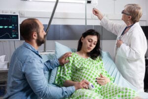 A pregnant woman in a hospital bed receiving an IV drip with a nurse monitoring her and holding a tablet.